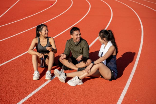 Three people sitting and chatting on an outdoor track in athletic wear, enjoying a break under the sun.