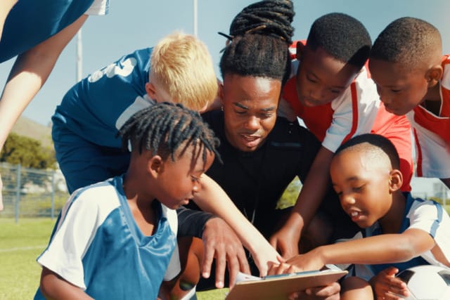 Coach and young soccer players reviewing a game plan on a clipboard outdoors on a sunny day.