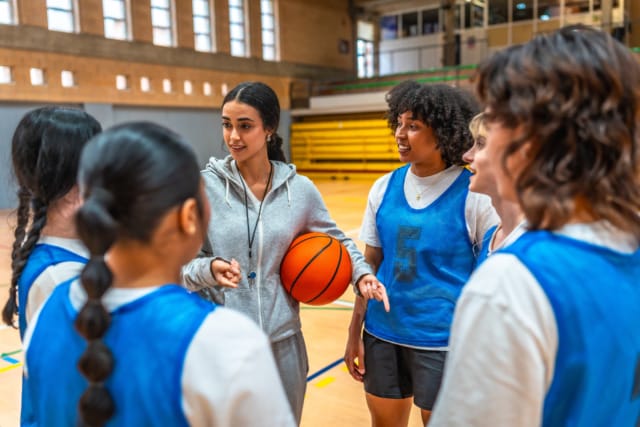 Coach discussing strategy with a women's basketball team in a gymnasium.