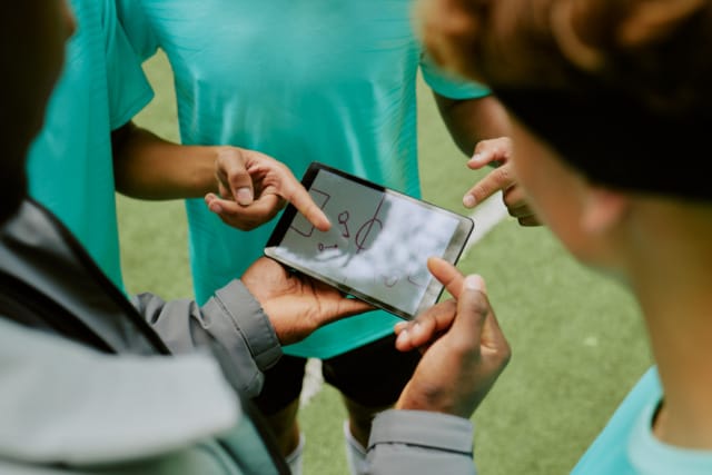 Soccer players strategizing with a coach using a tablet on the field, discussing game tactics and formations.