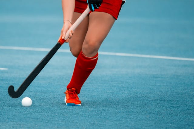 Field hockey player in red uniform dribbling a white ball on a blue field.