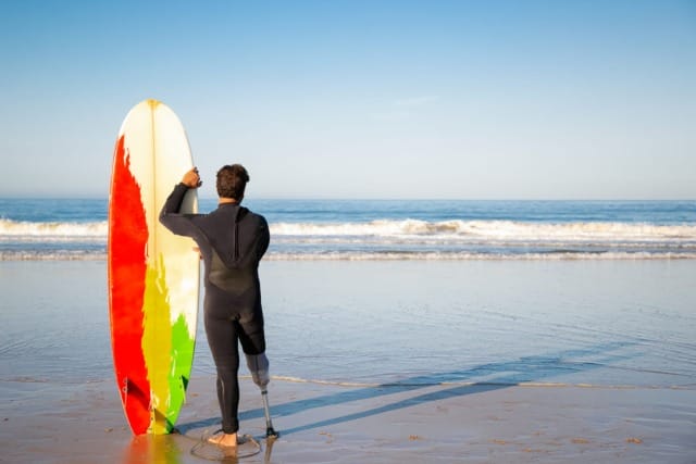 Surfer with prosthetic leg standing on beach holding colorful surfboard, looking at ocean waves under clear blue sky.