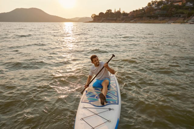 Man paddling on a stand-up paddleboard in a serene lake at sunset near a hilly coastline