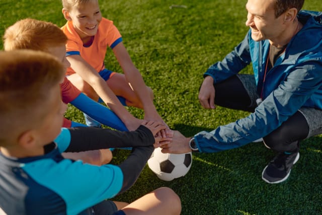 Coach and young soccer players sitting on grass in a huddle with hands stacked on a soccer ball, showing team spirit.
