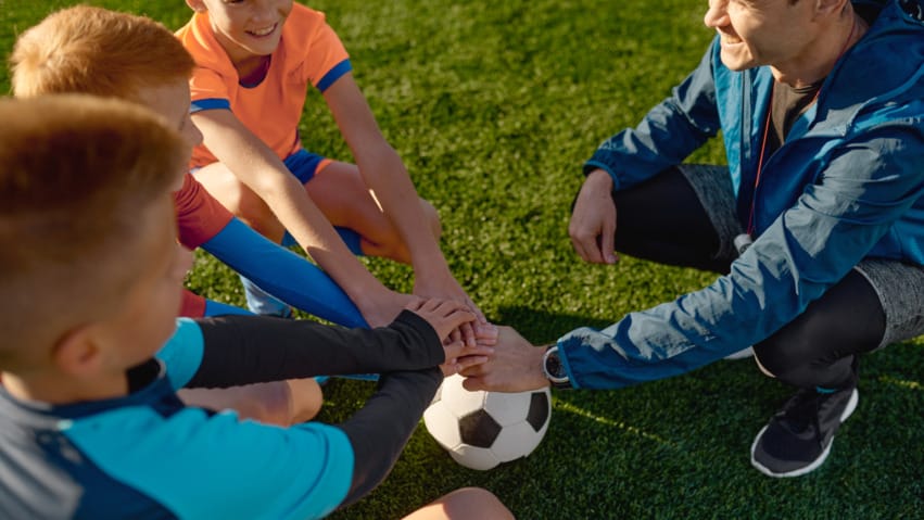 Coach and young soccer players sitting on grass in a huddle with hands stacked on a soccer ball, showing team spirit.