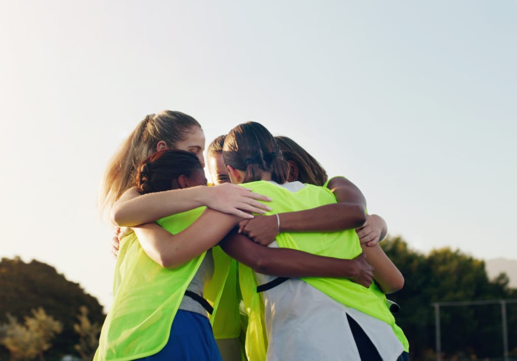 Group of young athletes in lime green jerseys huddling outside as a team, embracing each other during a sunny day.