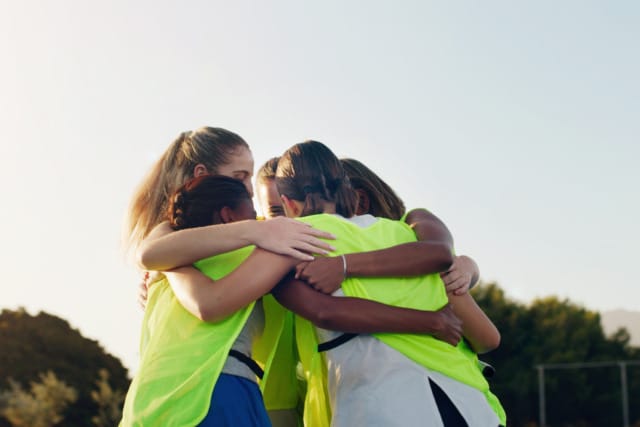 Group of young athletes in lime green jerseys huddling outside as a team, embracing each other during a sunny day.