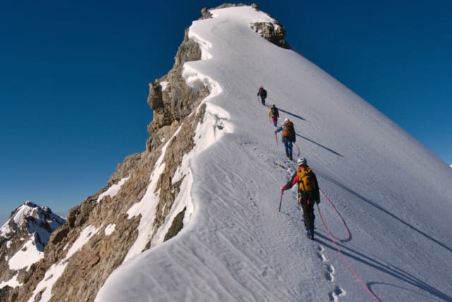 Mountaineers roped together, ascending a snowy ridge under a clear blue sky.