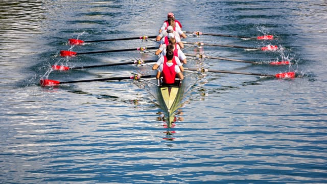 Rowing team in a synchronized motion on calm water, wearing red and white uniforms, with four rowers and a coxswain.