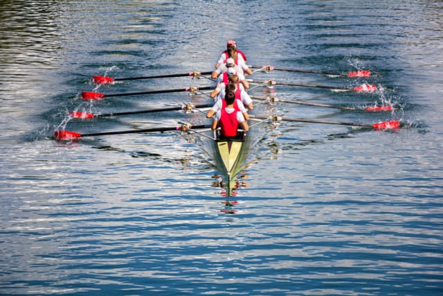 Rowing team in a synchronized motion on calm water, wearing red and white uniforms, with four rowers and a coxswain.