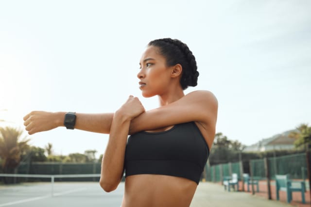 Woman stretching her arm on a tennis court, wearing workout gear and a smartwatch, with trees and a fence in the background.