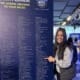 Person smiling and pointing at a list of charity runner names on a large display board at NYC Half event.