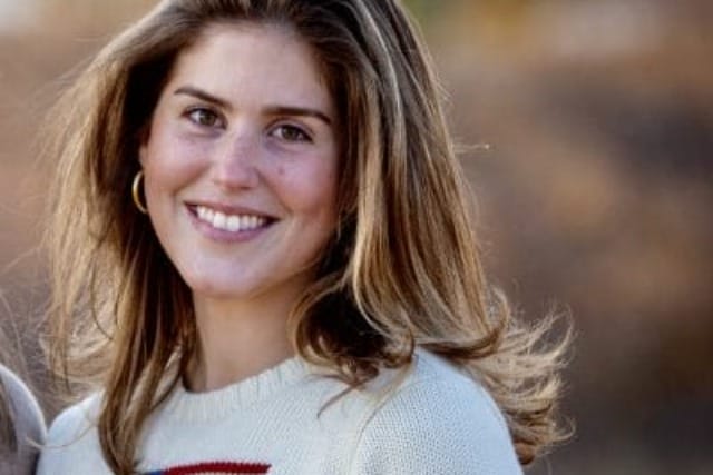 Smiling woman with long hair wearing a sweater featuring an American flag design, standing in an outdoor setting.