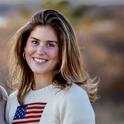 Smiling woman with long hair wearing a sweater featuring an American flag design, standing in an outdoor setting.