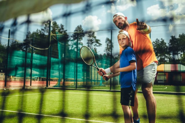 Coach teaching young boy tennis on an outdoor court, both holding rackets, net in foreground, sunny day.
