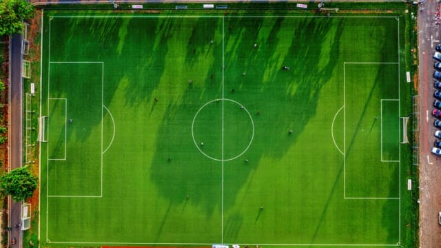Aerial view of a green soccer field with visible boundary lines, players, and surrounding trees and parking lot.