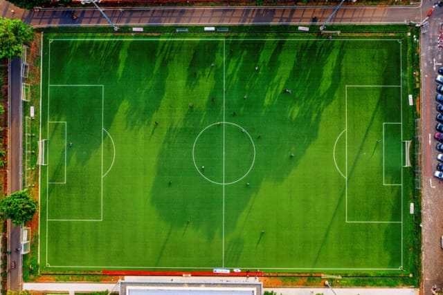 Aerial view of a green soccer field with visible boundary lines, players, and surrounding trees and parking lot.