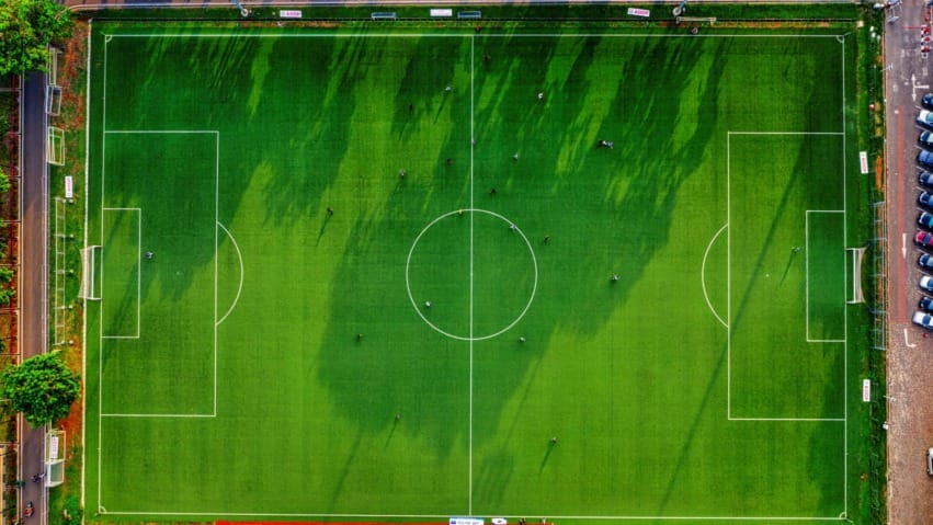 Aerial view of a green soccer field with visible boundary lines, players, and surrounding trees and parking lot.