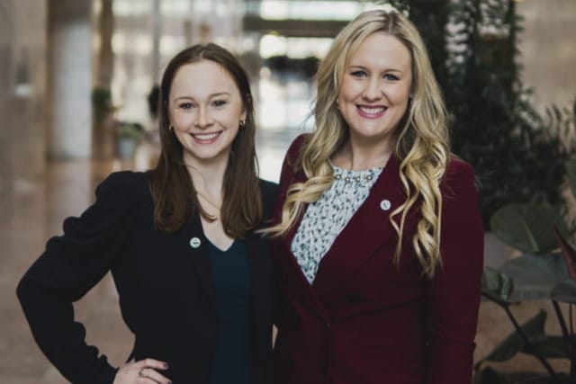 Two women smiling in business attire standing in a modern office lobby.