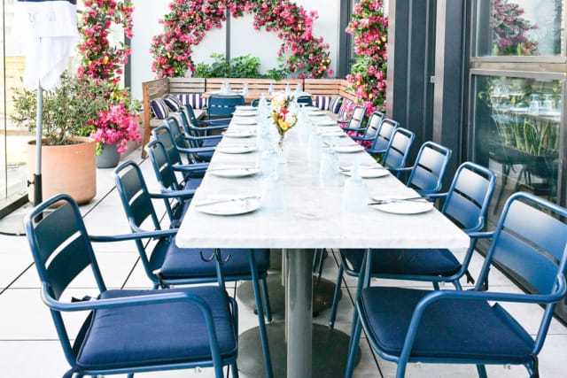 Outdoor rooftop dining setup with a long table, blue chairs, and pink floral decorations in the background.