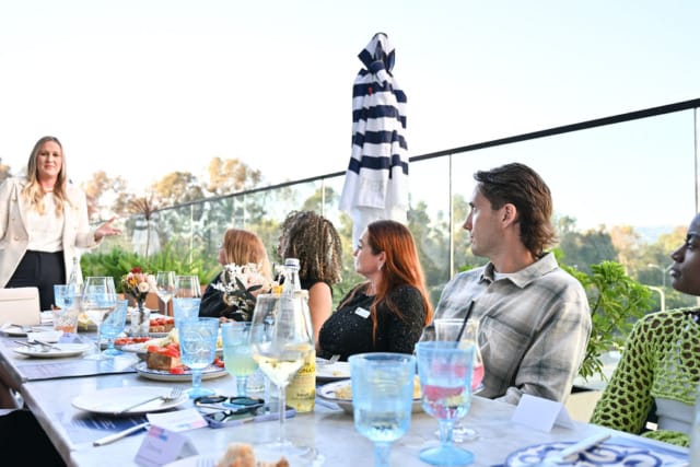 Group of people seated at an outdoor dining table, listening to a presenter, with a scenic view in the background.