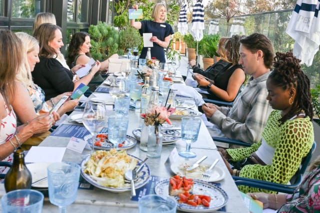 People seated around an outdoor dining table with food and drinks, engaged in a presentation by a standing woman.