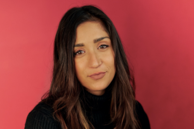 person with long hair wearing a black sweater poses against a red background