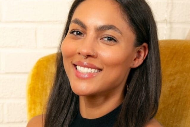 Person with long dark hair and a nose ring smiling against a yellow chair and white brick wall background.