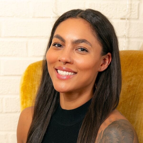 Person with long dark hair and a nose ring smiling against a yellow chair and white brick wall background.