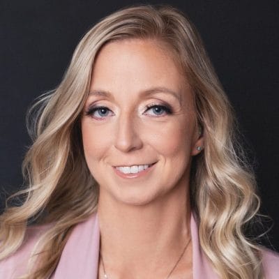 Smiling woman with long blonde hair wearing a pink blazer against a dark background.