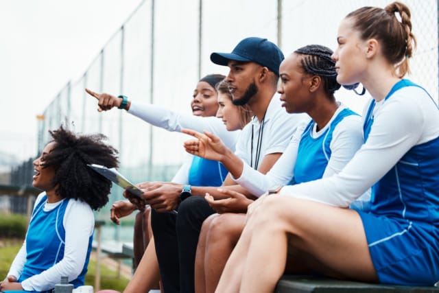 Basketball coach and team in blue uniforms sit on bench, strategizing and pointing on an outdoor court.