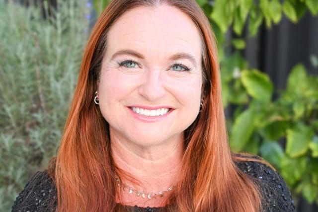 A woman with long red hair smiling and wearing a black top, sitting outdoors with greenery in the background.