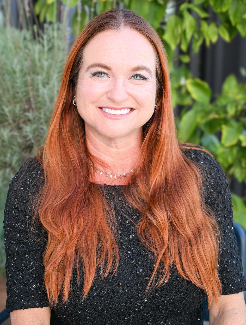 A woman with long red hair smiling and wearing a black top, sitting outdoors with greenery in the background.