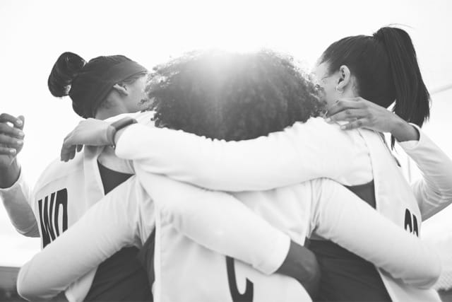 Sports team huddling with arms around each other in unity against a bright sky background.