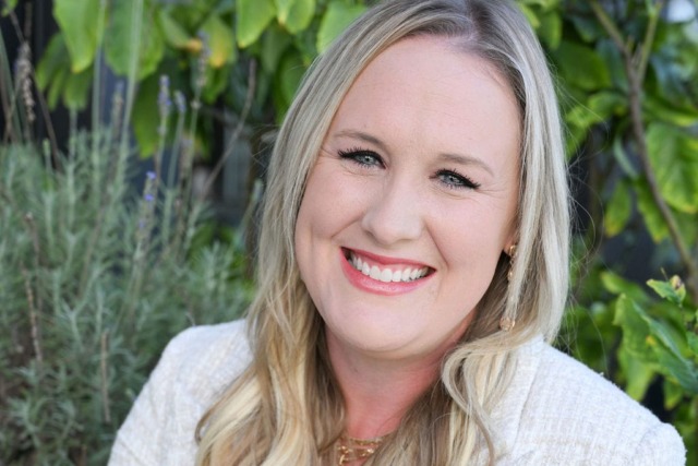 Smiling person with long blonde hair and a white blazer, sitting outdoors with green plants in the background.