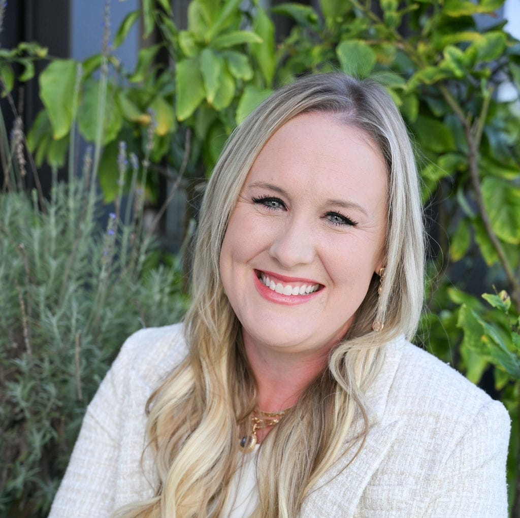Smiling person with long blonde hair and a white blazer, sitting outdoors with green plants in the background.