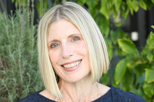Smiling woman with short blonde hair wearing a navy blue dress in front of green foliage.