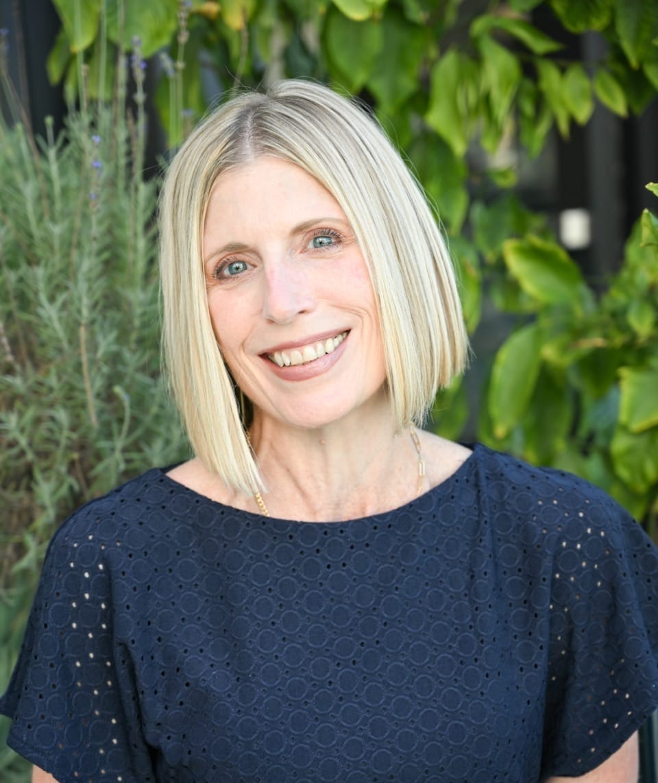Smiling woman with short blonde hair wearing a navy blue dress in front of green foliage.