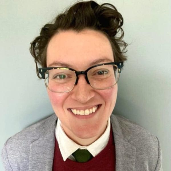 Person smiling warmly, wearing glasses, a gray blazer, red sweater, white shirt, and green tie against a light background.