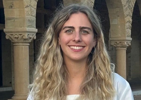 Person with long hair smiling, wearing a white coat, standing in front of stone arches.