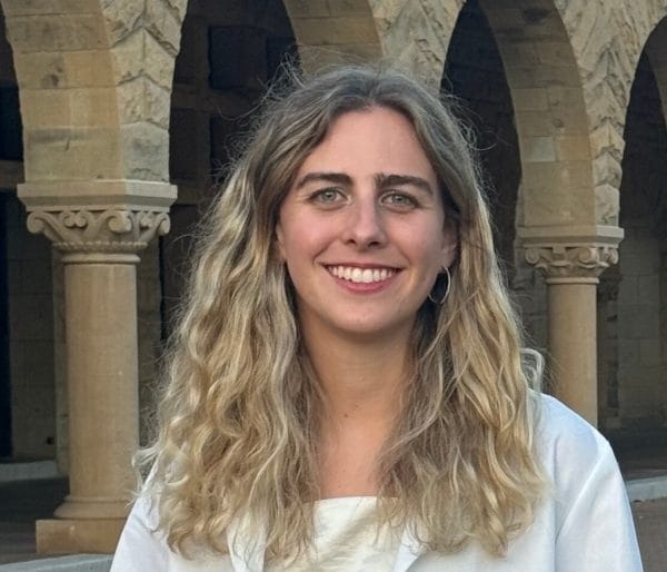Person with long hair smiling, wearing a white coat, standing in front of stone arches.