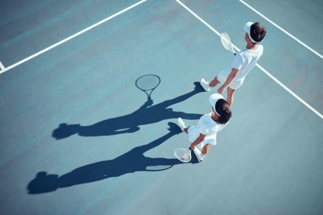 Two tennis players in white outfits and visors walking on a blue tennis court casting long shadows.