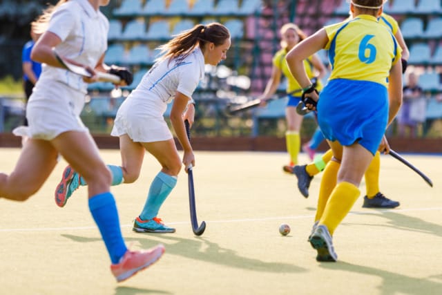 Women playing field hockey in action on a sunny day, wearing colorful uniforms on a green turf field.