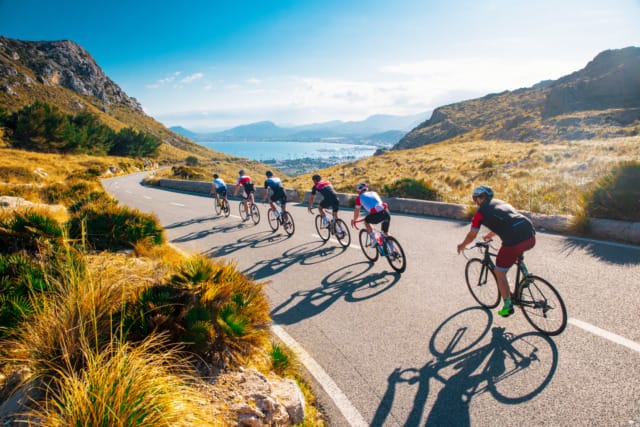 Cyclists riding on a scenic winding road with hills and a view of the ocean in the background.