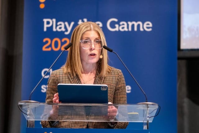 A woman speaking at a podium with a tablet in hand, in front of a "Play the Game 2023" banner.