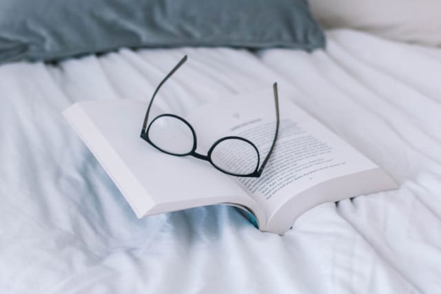 Open book with glasses resting on its pages, set on a white bedspread.