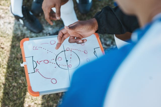 Soccer coach explaining strategy to players using a whiteboard outdoors on a field.