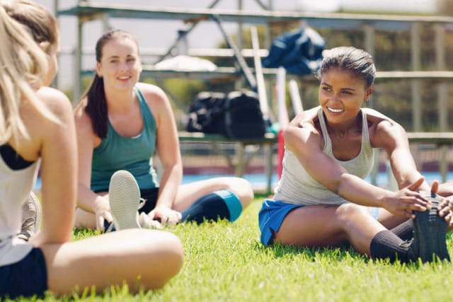 Three women sitting on grass, stretching and smiling during an outdoor workout session on a sunny day.