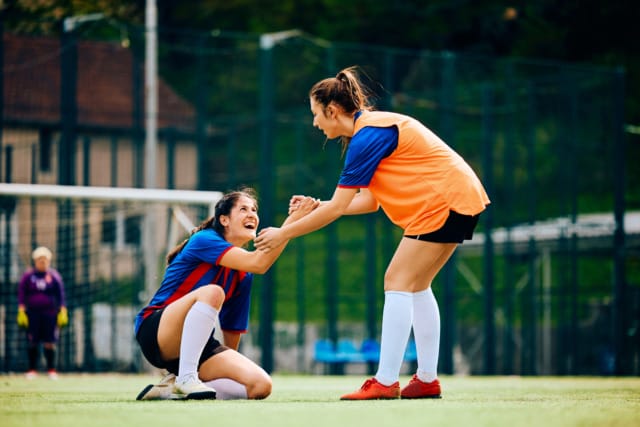 Soccer player in orange pinnie helps teammate in blue jersey off the ground on a soccer field during daytime.