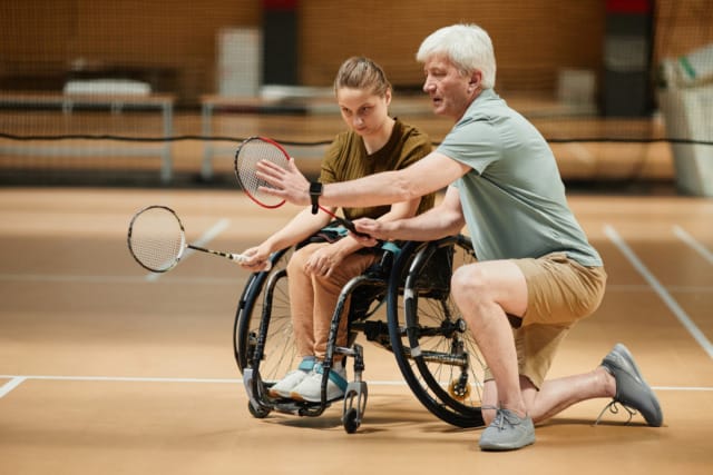 Coach instructing young wheelchair tennis player on court, both holding rackets, focus on technique and guidance.